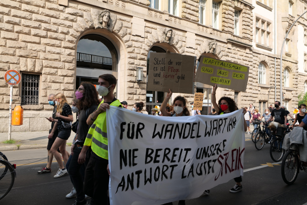 Eine Gruppe von Menschen marschiert auf einer Straße bei einer Demonstration, einige halten Schilder und andere fahren Fahrräder, im Hintergrund ein historisches Gebäude mit Rundbogenfenstern, Pfeilern und Skulpturen.
