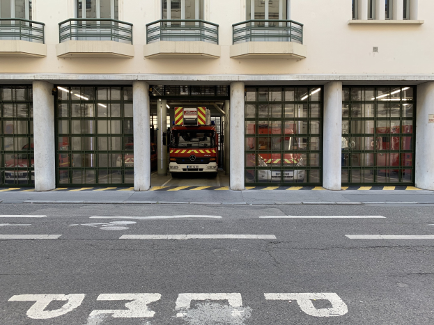 Feuerwache mit einem vor der Tür geparkten Feuerwehrauto, das Fenster, Geländer, Säulen, Glastüren und eine Straße am Boden zeigt.