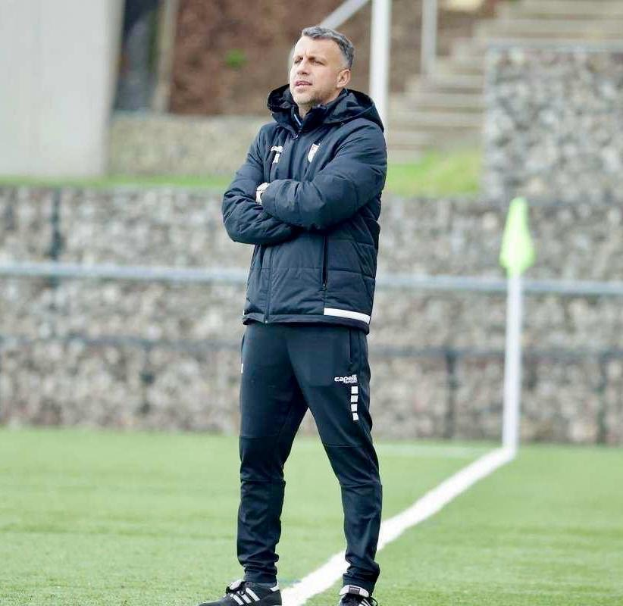 Ein Füßballtrainer mit überkreuzten Armen auf einem Feld, der eine schwarze Jacke und Hose trägt, vor einem Zaun, einer Treppe und einer Wand.
