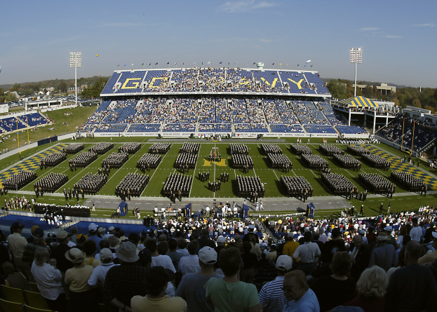Eine große Menschenmenge schaut sich ein Fußballspiel in einem Rasenstadion unter einem klaren blauen Himmel an, mit Flutlichtmasten, Bäumen und Gebäuden im Hintergrund.