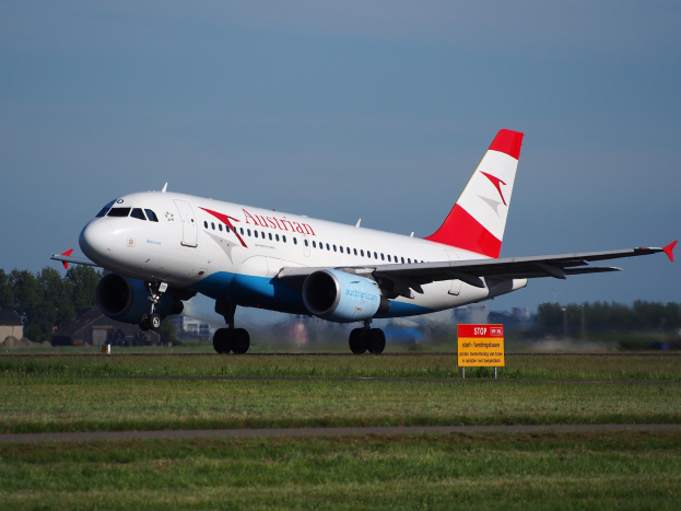 Austrian Airlines Airbus A320-200 beim Start am Frankfurt Airport mit einer Tafel im Vordergrund, Gras, Bäumen, Gebäuden und einem klaren blauen Himmel im Hintergrund.