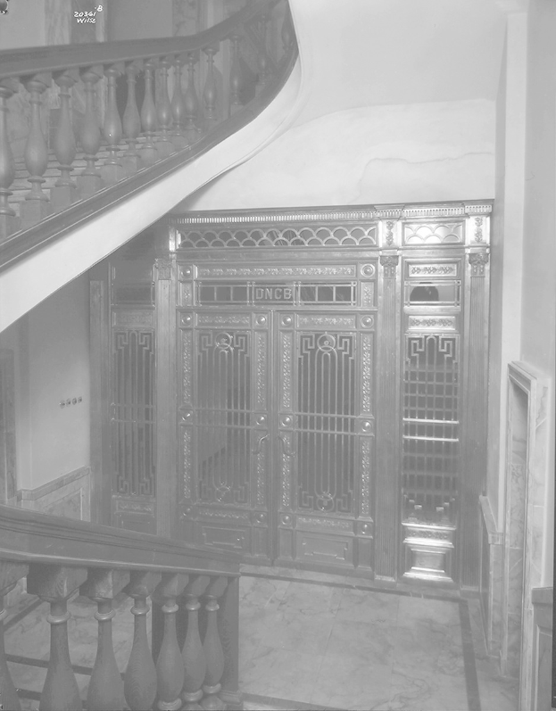 Schwarzes und weißes Foto einer Treppe mit einem linken Geländer, einer Wand im Hintergrund, einem Boden unten und einer Tür in der Mitte, die wie ein Safe aussieht.