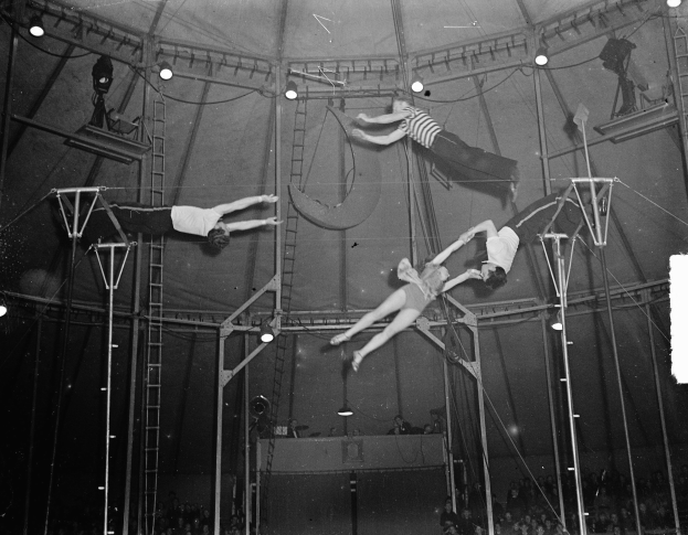 Schwarzes und weißes Foto von Akrobaten, die in einem Zirkuszelt auftreten, mit einer Zuschauermenge darunter.