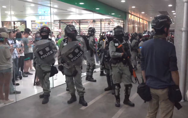 Eine Gruppe von Polizisten steht vor einer Menschenmenge, einige tragen Helme und halten Waffen, in einem Einkaufszentrum mit einer Säule auf der rechten Seite, einer Glaswand im Hintergrund und einer beleuchteten Decke.