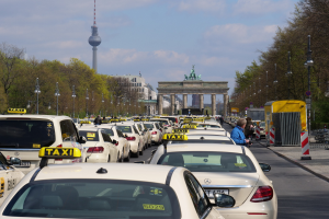 Eine belebte Berliner Straße mit parkenden Taxis, Fußgängern auf dem Gehweg, Laternen, Bäumen, Gebäuden und einem fernen Bogen mit Statuen und Turm unter einem bewölkten Himmel.