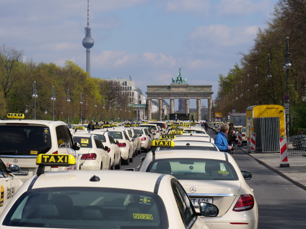 Eine belebte Berliner Straße mit parkenden Taxis, Fußgängern auf dem Gehweg, Laternen, Bäumen, Gebäuden und einem fernen Bogen mit Statuen und Turm unter einem bewölkten Himmel.