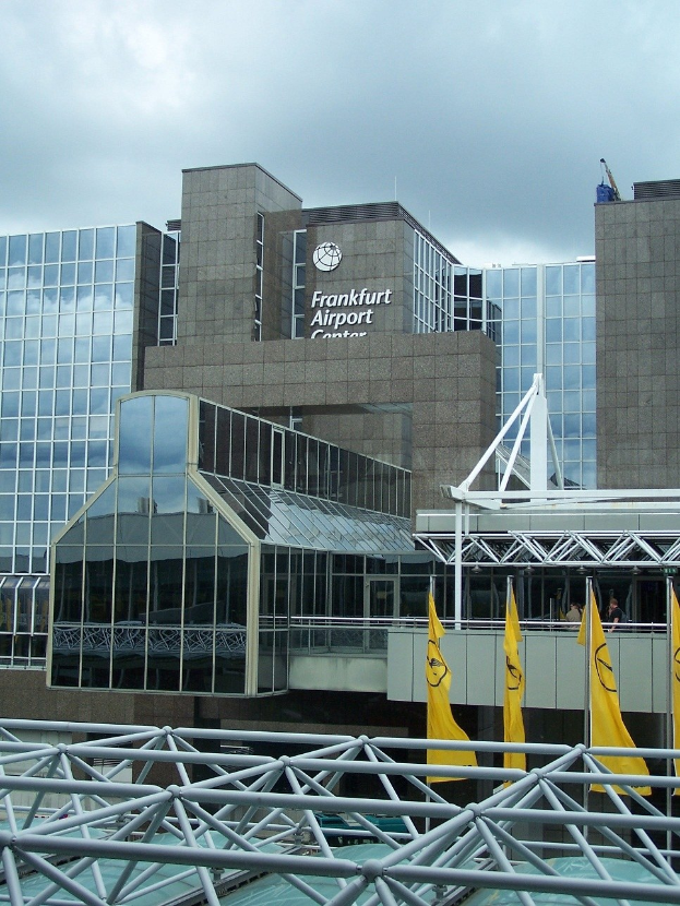 Frankfurt Airport Gebäude mit Glaswänden, Text, gelben Flaggen und Eisenstangen unter einem bewölkten Himmel.