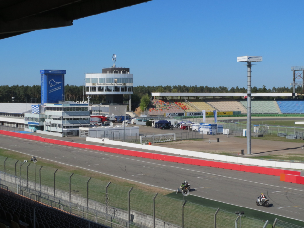 Gruppe von Motorradfahrern auf einer Rennstrecke, umgeben von einem Metallzaun, Gras, Fahrzeugen, einem Gebäude, einem Schild, Pfosten, einem Stadion, einem Dach mit Deckenleuchten, Bäumen und einem bewölkten Himmel.
