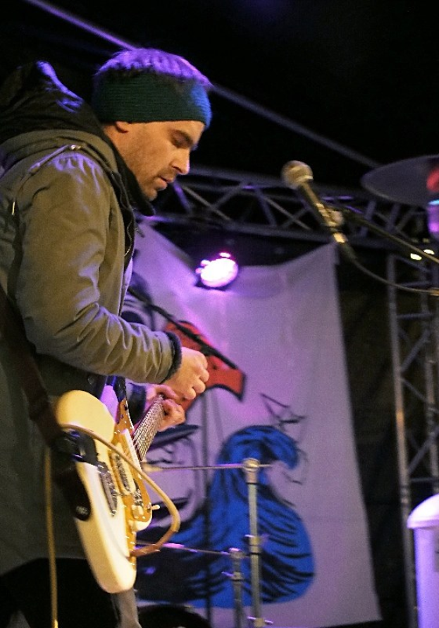 Ein Mann in einer Jacke und einer Mütze spielt eine E-Gitarre auf der Bühne mit einem Mikrofon vor sich, einem Schlagzeug zu seiner Rechten und beleuchteten Bannern im Hintergrund.