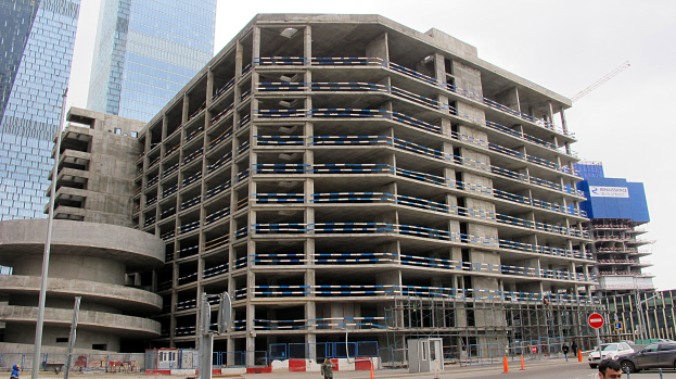 Großes Bauprojekt in einer städtischen Umgebung mit Wolkenkratzern, Fahrzeugen, Fußgängern, Verkehrsinfrastruktur und Kränen vor einem klaren Himmel.