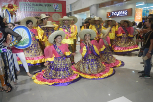 Eine Gruppe von Menschen in bunten Kleidern und Hüten tanzt in einem Einkaufszentrum, während andere Musikinstrumente halten und ein Banner tragen, während einer festlichen Cinco de Mayo-Feier.