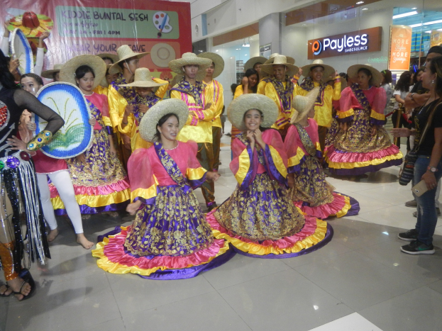 Eine Gruppe von Menschen in bunten Kleidern und Hüten tanzt in einem Einkaufszentrum, während andere Musikinstrumente halten und ein Banner tragen, während einer festlichen Cinco de Mayo-Feier.
