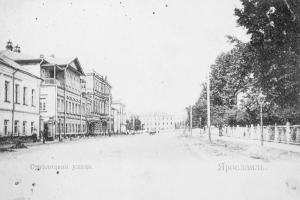 Ein Schwarz-Weiß-Foto einer Stadtstraße mit Gebäuden, Bäumen, Pfählen, Lampen und einem Zaun auf der rechten Seite, mit Text unten.
