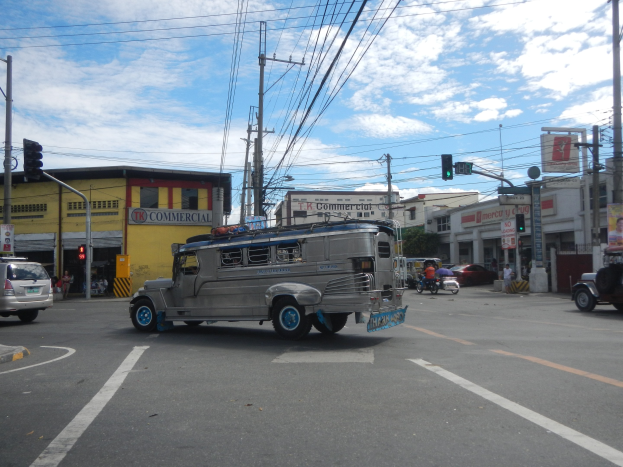Eine Stadtstraße mit einem Jeep, der an einer Ampel vorbeifährt, umgeben von Fahrzeugen, Strommasten, Gebäuden und einem bewölkten Himmel.