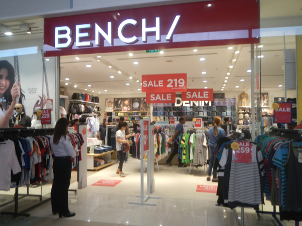 Ein Bekleidungsgeschäft namens 'Bench' in einem Einkaufszentrum, das Kleidung an Bügeln zeigt, mit beleuchteter Decke, Informationsschildern und einem Bildschirm auf der linken Seite, während Menschen herumlaufen.