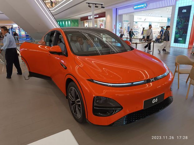 Oranges Elektroauto in einer Ausstellungshalle mit einer Gruppe von Menschen drumherum, umgeben von Stühlen, einem Wagen, Geschäften, Namensschildern und Lichtern im Hintergrund.
