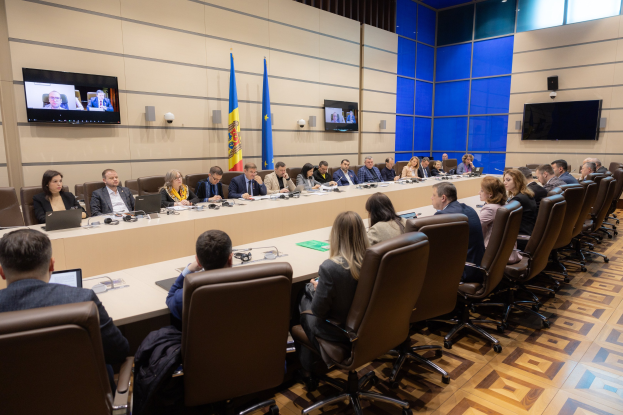 Große Gruppe von Menschen an einem langen Tisch mit Laptops, Mikrofonen und Papieren während einer Sitzung des Europäischen Rates für Menschenrechte, mit drei Flaggen und zwei Fernsehgeräten im Hintergrund.