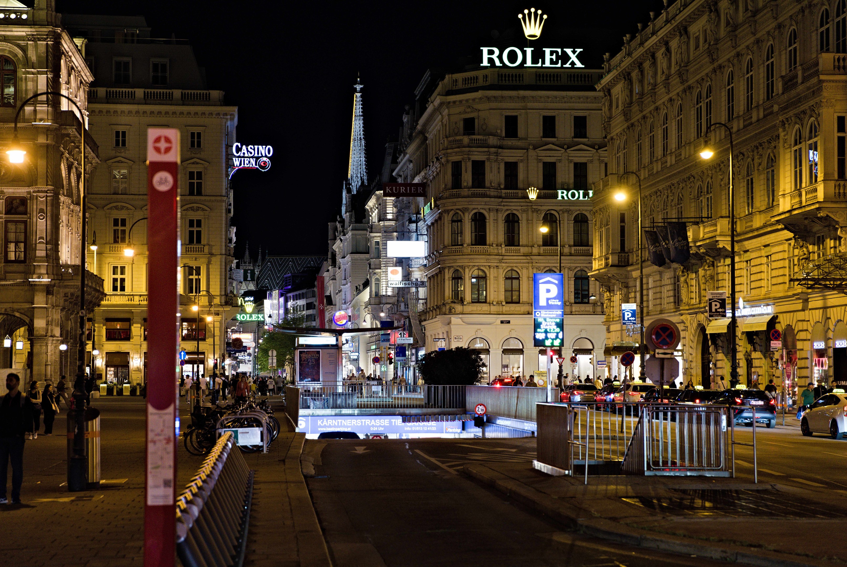 Eine nächtliche Stadtstraße mit fahrenden Autos, Fußgängern, parkenden Fahrrädern, Straßenlaternen, hohen Gebäuden, Absperrungen, Polen und einem Rolex-Logo im Hintergrund.