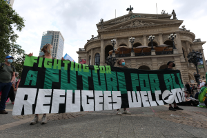 Eine Gruppe von Menschen mit Masken hält ein Banner mit der Aufschrift "Kämpfen für eine Zukunft für alle Flüchtlinge willkommen" vor einem Gebäude mit Laternenmasten, Bäumen und einem bewölkten Himmel.