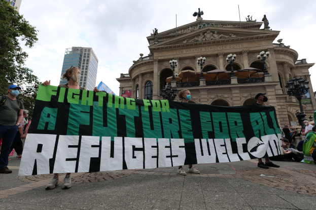 Eine Gruppe von Menschen mit Masken hält ein Banner mit der Aufschrift "Kämpfen für eine Zukunft für alle Flüchtlinge willkommen" vor einem Gebäude mit Laternenmasten, Bäumen und einem bewölkten Himmel.