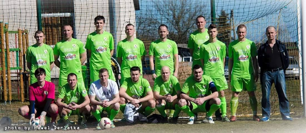 Eine Gruppe von Männern in grünen T-Shirts steht auf einem Fußballfeld, einige in Hockstellung, mit einem Ball davor und einem Torpfosten rechts daneben, vor einem Hintergrund aus Bäumen, Gebäuden und Fahrzeugen.