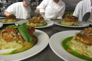 Drei Köche in einer Küche mit Mützen und Handschuhen bereiten Essen auf Tellern vor, die mit verschiedenen Gegenständen gefüllt sind, mit Regalen voller Vorräte im Hintergrund.