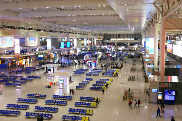 Innenansicht eines Flughafen-Terminals mit Menschen, blauen Stühlen, Geschäften, Informationsanzeigen, Bildschirmen, tragenden Säulen, Deckenbeleuchtung und Stützstangen.