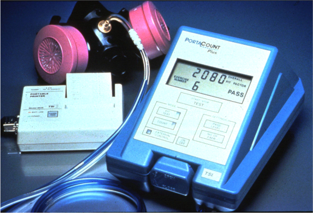 Eine Gasmaske wird auf einen Tisch neben ein elektronisches Gasdetektorgerät mit einem angeschlossenen Kabel gelegt.