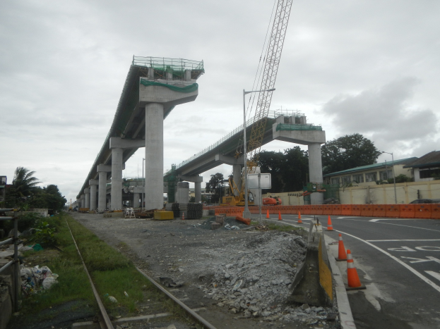 Baustelle mit einer Brücke im Hintergrund, Straße mit Verkehrskegeln markiert, Bahnschiene links, verstreute Steine und Gras, Bäume und Gebäude auf beiden Seiten und ein bewölkter Himmel.