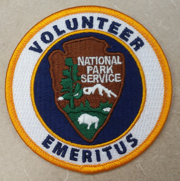 Ein rundes Patch mit einem blauen Rand, weißem Hintergrund und fetter schwarzer Schrift, die "Volunteer National Park Service Emeritus" in der Mitte eines gelben Kreises liest.