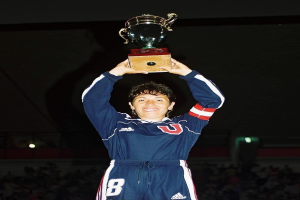 Eine Frau in einem blauen Kleid hält einen Pokal hoch und lächelt, umgeben von einer jubelnden Menge in einem hell erleuchteten Stadion.