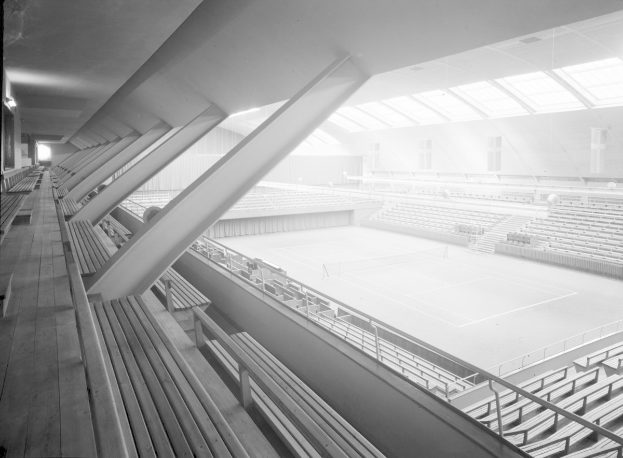 Ein Schwarz-Weiß-Foto eines Indoor-Tennisplatzes in einem Stadion mit Sitzreihen, einem Geländer und Deckenlampen.