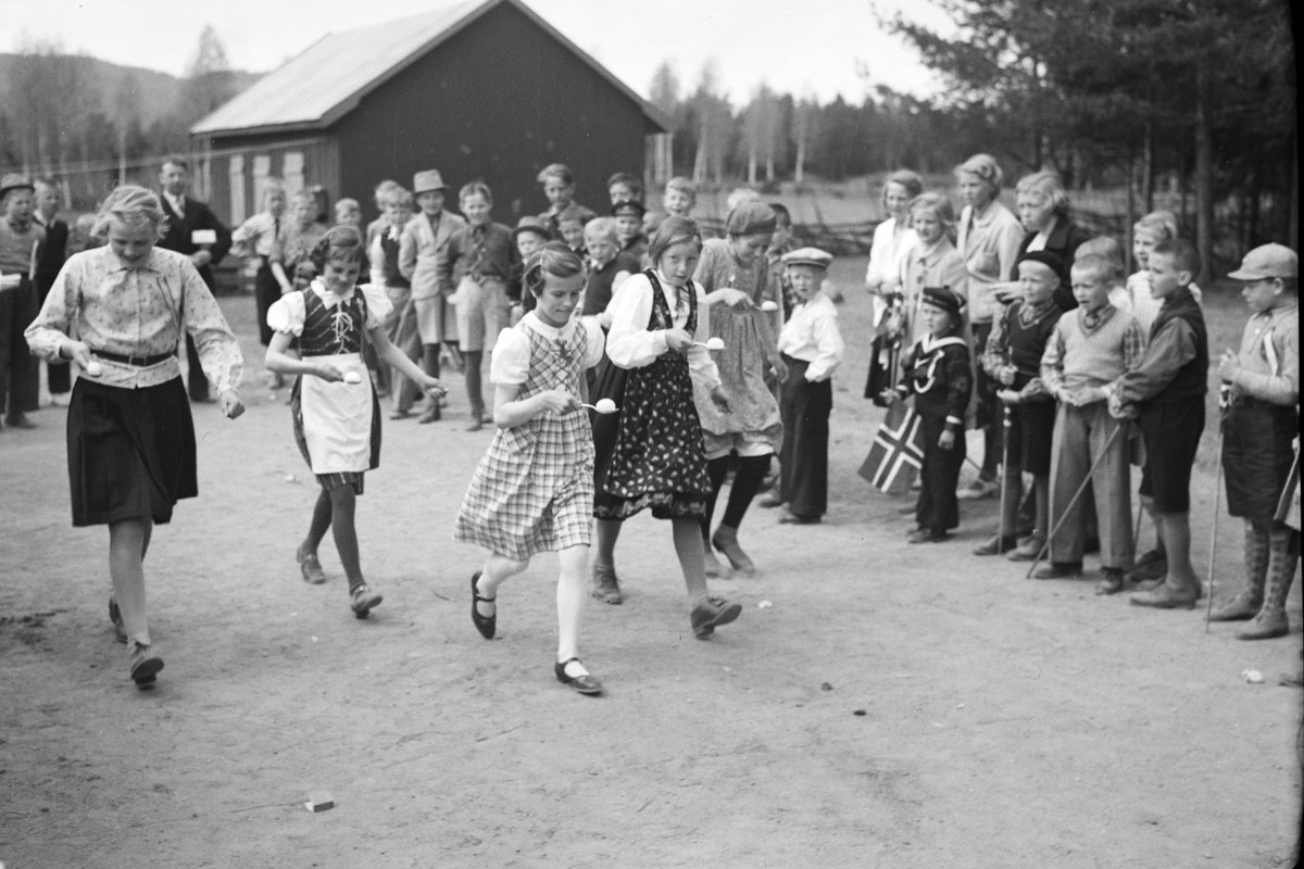 Schwarzes Foto von Kindern, die vor einer Menge mit Fahnen Fußball spielen, mit Bäumen und einem Haus im Hintergrund.