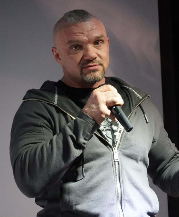 Ein Mann in einer schwarzen Kapuzenjacke und einem schwarzen Anzug mit einem weißen Hemd, glattem Haar und einem selbstbewussten Gesichtsausdruck hält ein Mikrofon, waßrend er vor einer Wand steht.