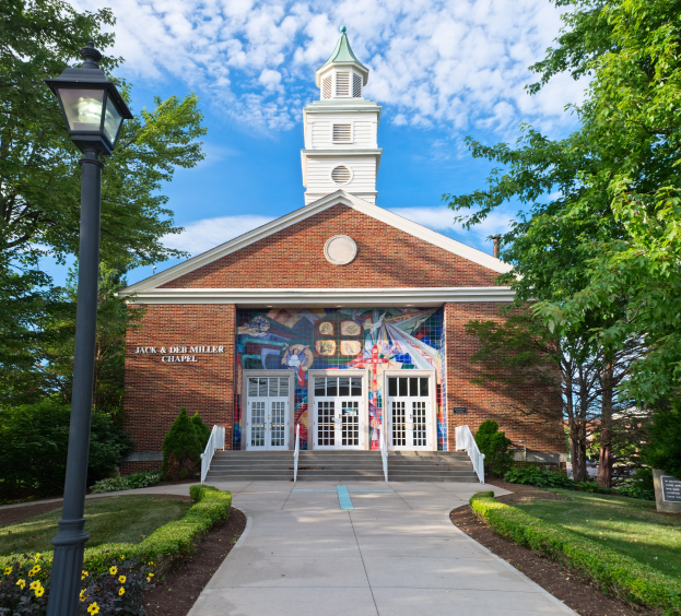 Die Jackson & De Miller Chapel, ein Backsteingebäude mit einem zentralen Glockenturm, umgeben von Fenstern, Türen, Treppen, Geländern, Pflanzen, Gras, einem Weg, einem Straßenmast, einer Baumgruppe und unter einem bewölkten Himmel.