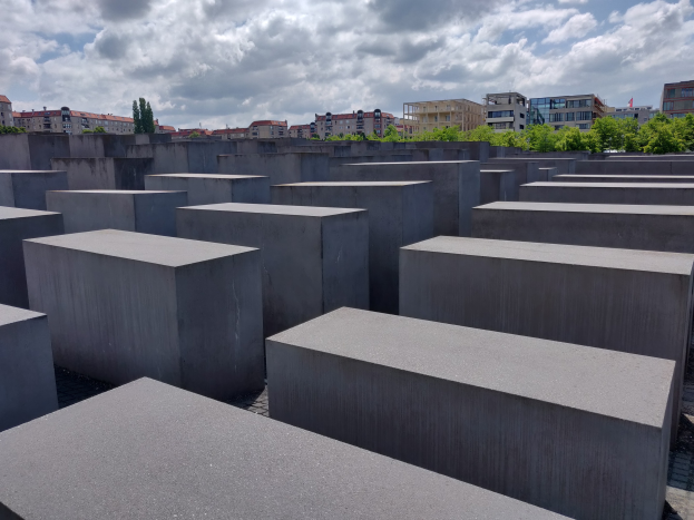 Luftaufnahme des Holocaust-Mahnmals für die ermordeten Juden Europas in Berlin, die ein Gittermuster aus Betonblöcken zeigt, mit Bäumen, Gebäuden und Wolken im Hintergrund.