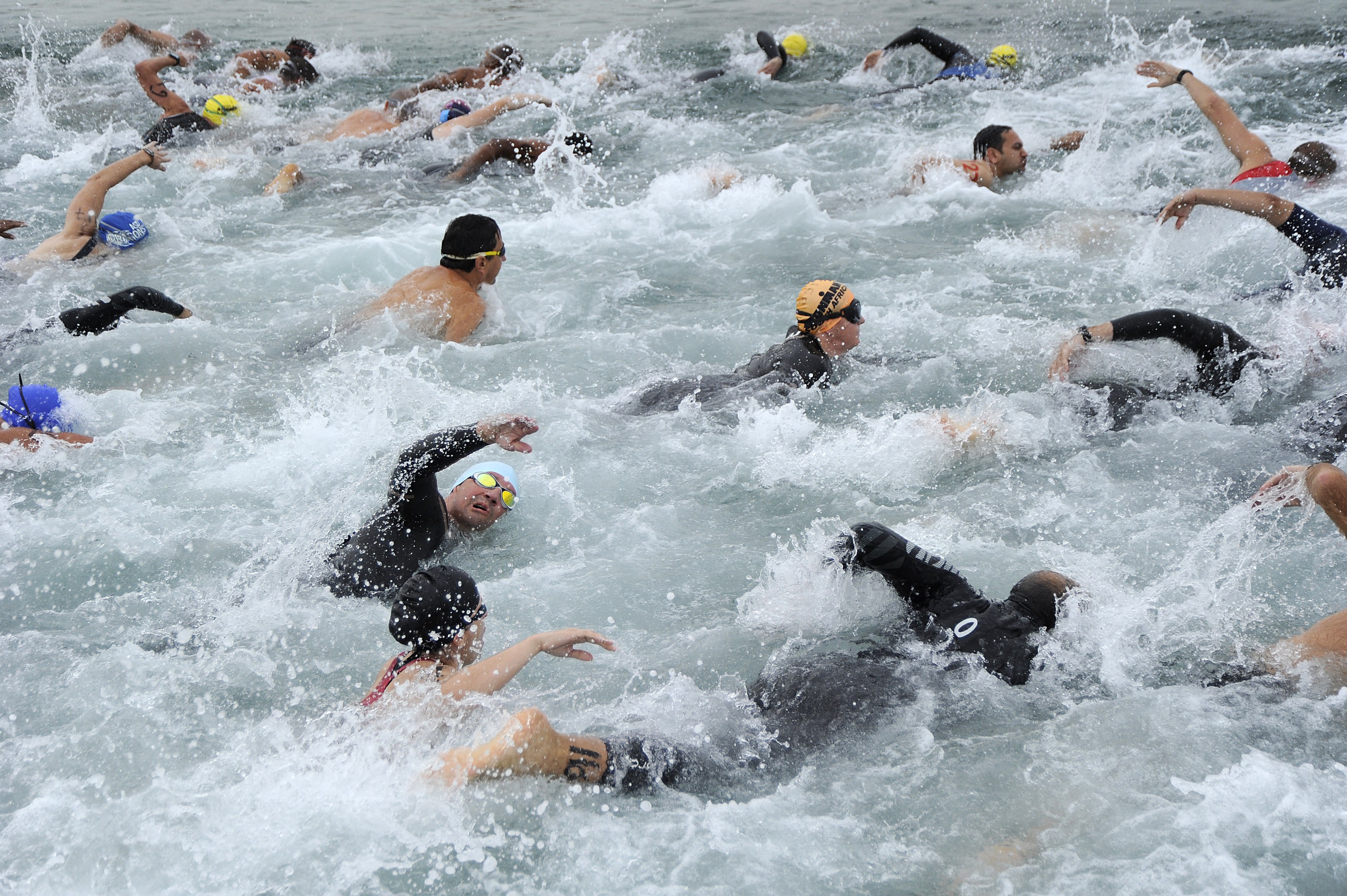 Gruppe von Triathleten, die in tiefem Blauwasser während eines Rennens schwimmen, tragen Neoprenanzüge, Schwimmkappen und Schwimmbrillen, mit ausgestreckten Armen in Richtung der Ziellinie unter strahlendem Sonnenlicht.
