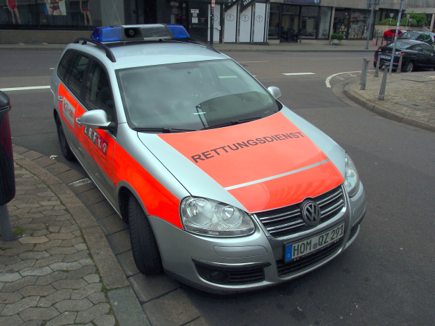 Polizeiauto am Straßenrand mit Mülltonne auf dem Gehweg links, Gebäude und Bäume im Hintergrund.