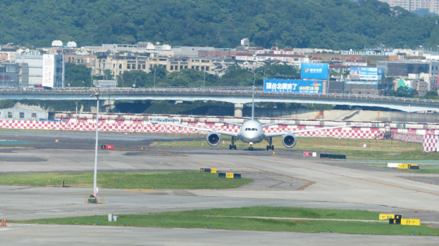 Flugzeug startet von einer Flughafenrollbahn mit umgebenen Infrastruktur- und Natürelementen unter einem klaren blauen Himmel.