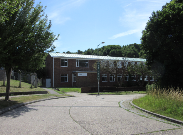 Ein Schulgebäude mit Fenstern, einem Straßenschild, einer Tafel, einer Baumgruppe, Gras, einem Zaun und einem bewölkten Himmel, das inmitten eines Parks mit saftigem Gras liegt.
