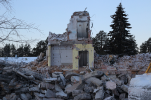 Ein abgerissenes Gebäude in einer verschneiten Landschaft, umgeben von Bäumen und einem klaren Himmel, mit Schutt und Trümmern drumherum.