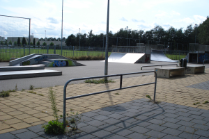 Ein Skatepark mit Rampen, Geländern, Grünflächen, Polen, Bäumen, Gebäuden und einem bewölkten Himmel im Hintergrund.