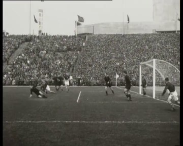 Schwarzes und weißes Foto des WM-Finals 1958 zwischen Manchester United und Liverpool, das Spieler auf dem Feld, ein Torpfosten rechts, Zuschauer in den Rängen und Fahnen im Hintergrund zeigt.
