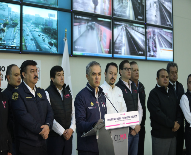 Gruppe von Männern, die neben einem Rednerpult mit einem Mikrofon und einer Tafel mit der Aufschrift "Mexikanische Zoll- und Grenzschutzbehörde" stehen, vor einer Wand mit Fernsehern und einer Flagge.