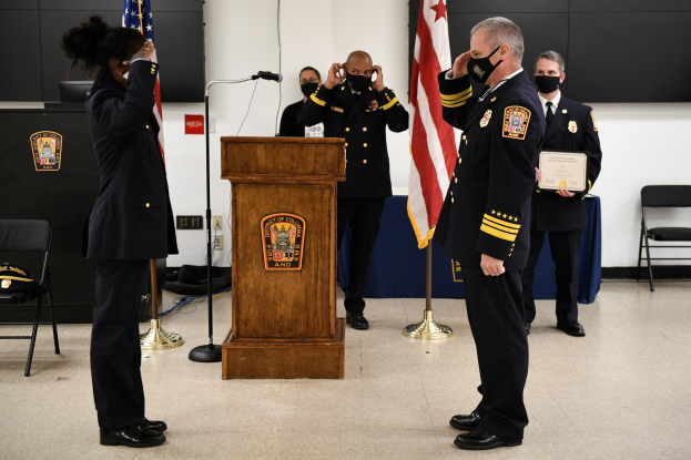 Gruppe uniformierter Personen in Masken vor einem Rednerpult mit Mikrofon während einer Vereidigung eines neuen Feuerwehrchefs, der Urkunden hält und von Fahnen, Stühlen, Tischen und einer Wand umgeben ist.
