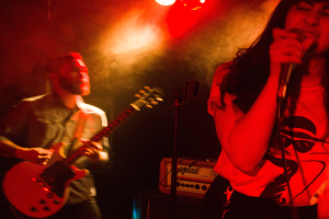 Eine Frau singt in ein Mikrofon auf der Bühne, während ein Mann eine akustische Gitarre im Hintergrund spielt, mit Deckenlampen, die die lebendige Kulisse beleuchten.