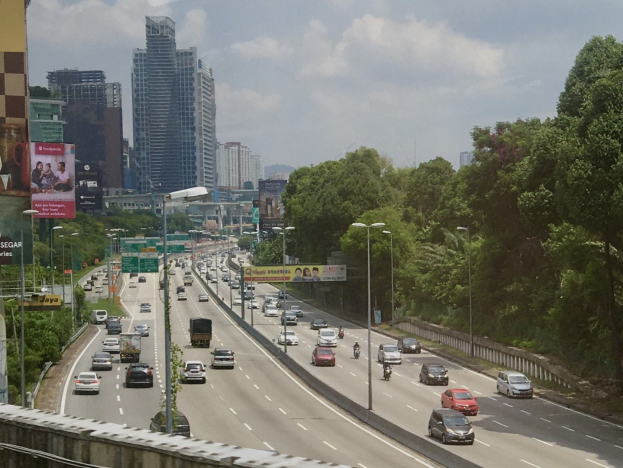 Eine belebte Autobahn mit hohen Gebäuden, Straßenlaternen, Schildern, Bäumen und einem bewölkten Himmel.