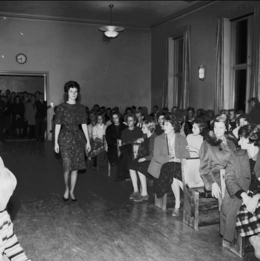 Eine Frau in einem schwarzen und weißen Kleid läuft über den Laufsteg, während das Publikum eine Modenschau zur Feier des Frauenwahlrechts betrachtet.