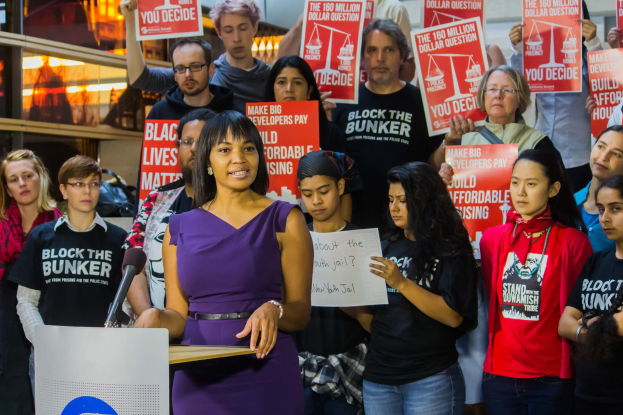Eine Black Lives Matter-Aktivistin spricht an einem Pult mit Mikrofon vor einer Menge mit Schildern, mit Bühnenbeleuchtung und Equipment im Hintergrund.