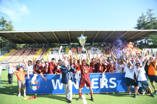 Eine Gruppe von Menschen auf einem Fußballfeld, einige halten einen Pokal, mit einem Banner, Mülltonne, verstreuten Kleidern, einem Zaun, einem Dach mit Metallstangen, sitzende Zuschauer unter dem Dach, Bäume und ein bewölkter Himmel.